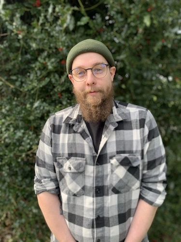 A Beared man stands infron of trees smiling